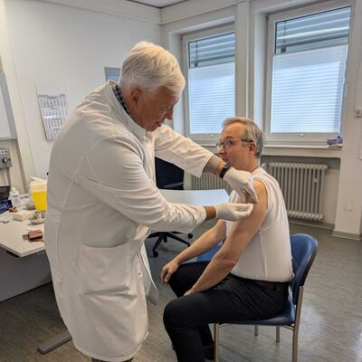 Dominik Meyer zu Schlochtern, Erster Kreisrat des Landkreises Gifhorn, sitzt auf einem Stuhl im Gesundheitsamt und erhält von Josef Kraft, Leiter des Gesundheitsamtes des Landkreises Gifhorn, die Grippeschutzimpfung in den Oberarm.